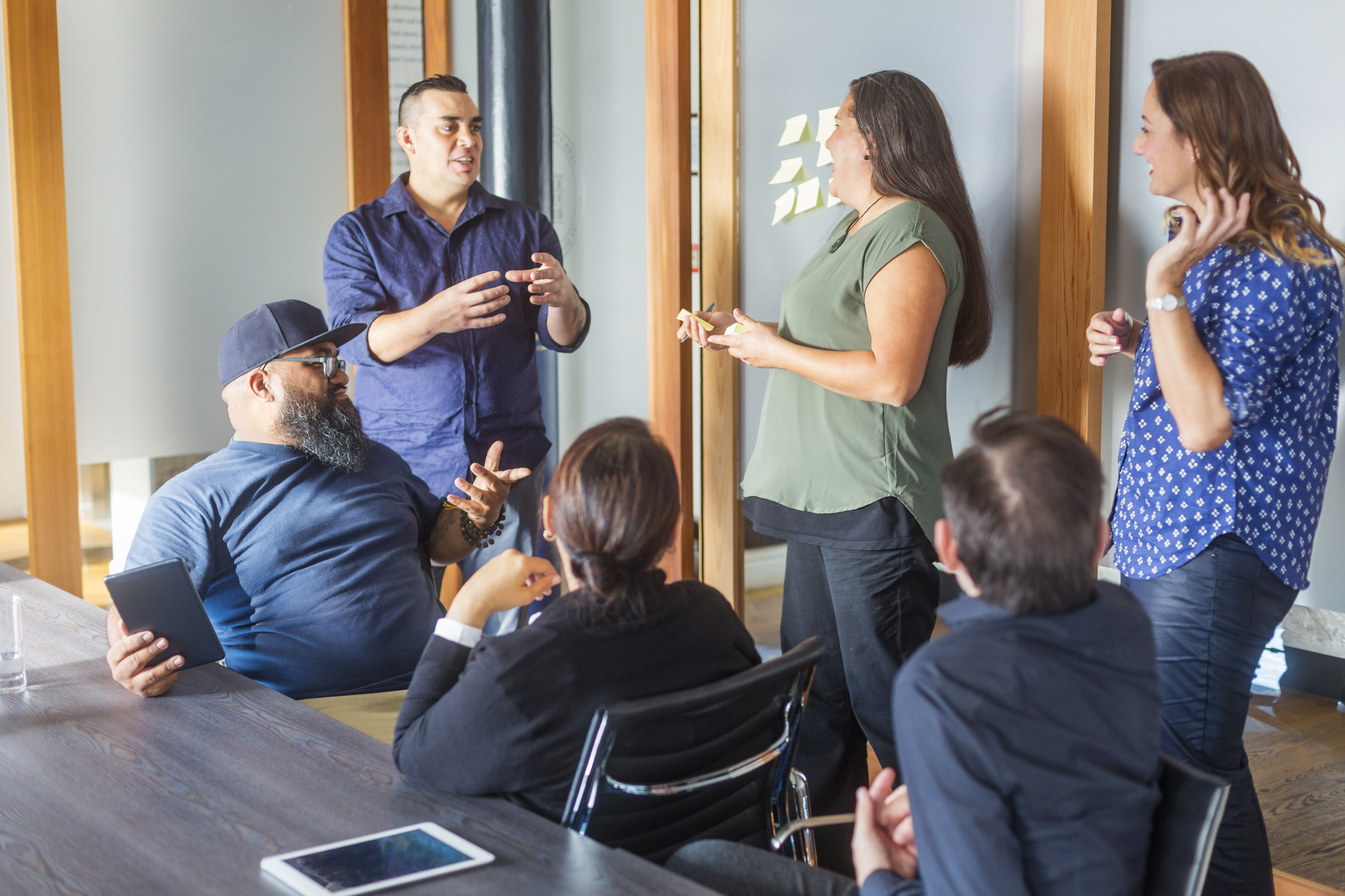 A group of six people in a conference room talking to each other. Three people are standing, from left a man in a blue shirt, a Māori woman wearing a green top and a woman wearing a patterned blue and white shirt all talking and laughing. Sitting in front of them at a table are, from left, a man wearing glasses and a cap, a woman with her hair in a bun wearing a black blazer and a man wearing a navy shirt.