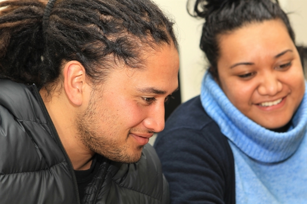 A young Māori man sitting next to a young Māori woman. The man has dreadlocks is wearing a black puffer jacket. The woman has dark hair in a bun and is wearing a blue knitted jumper. They are both smiling.